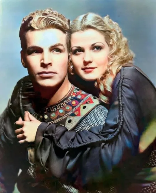 A vintage color photograph shows a man and woman in period costumes, the woman embracing the man from behind. The man has a…