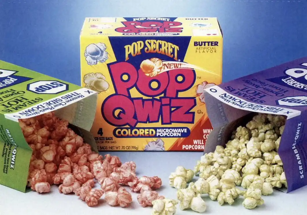 A vintage advertisement image shows a yellow box of Pop Secret Pop Qwiz colored microwave popcorn, flanked by two open bags…