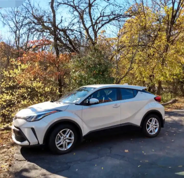 A white Toyota C-HR compact SUV is parked on a cracked asphalt road surrounded by autumn trees with yellow and orange…