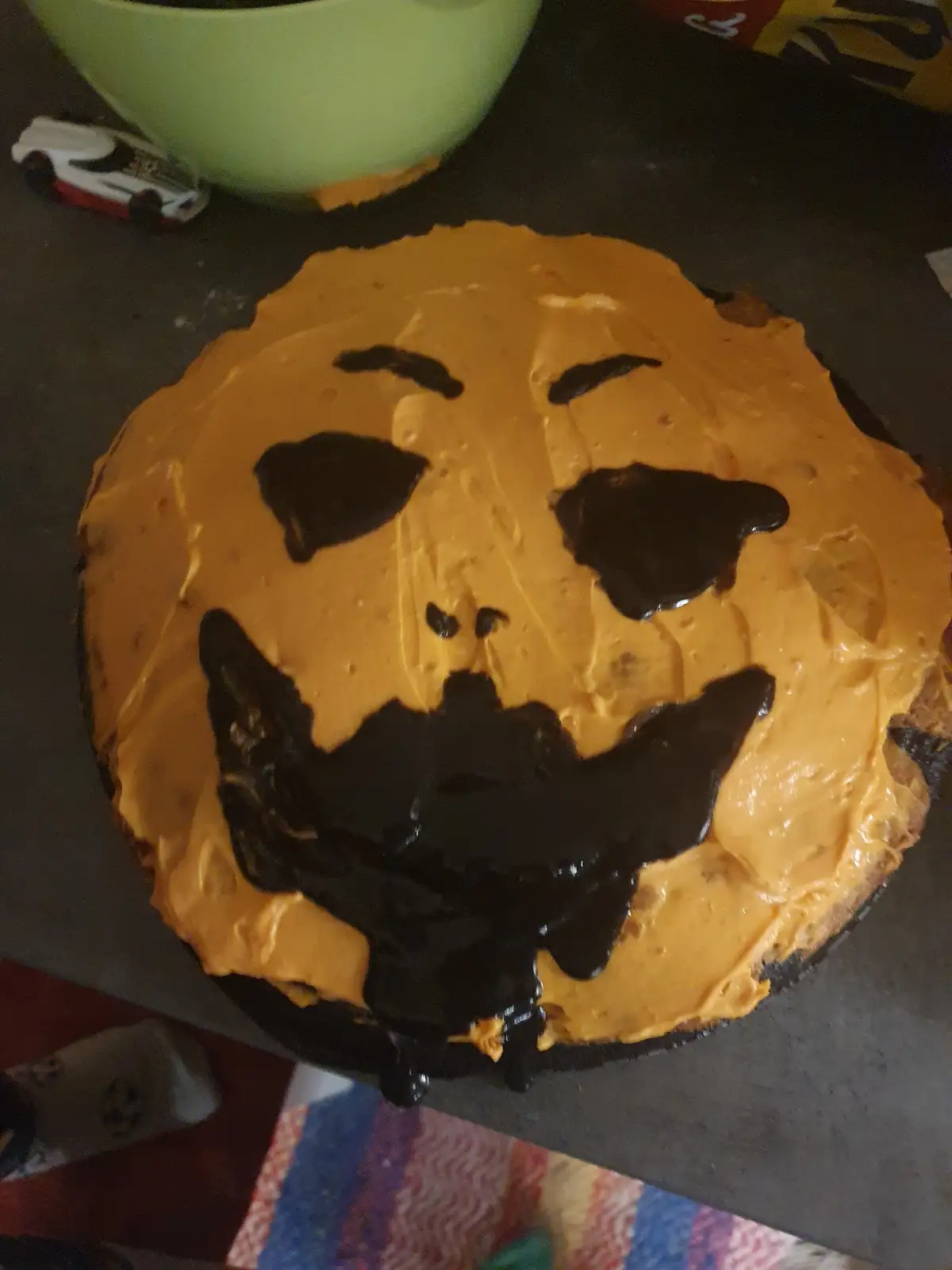 A round cake covered in orange frosting with crude black icing forming a jack-o'-lantern face, including eyes, nose, and a…