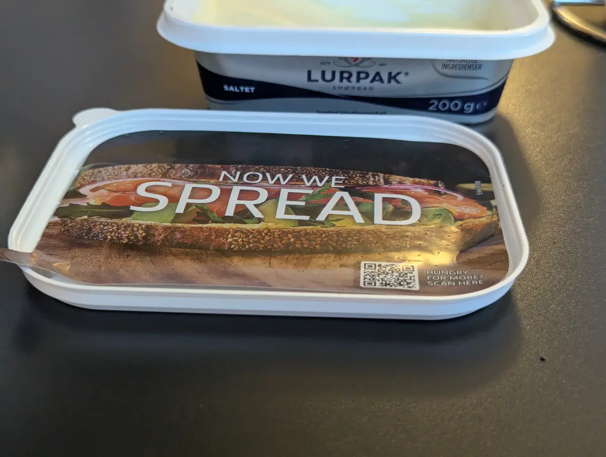A close-up photo shows a white plastic tub of Lurpak butter with a label that reads 'NOW WE SPREAD' over an image of a…
