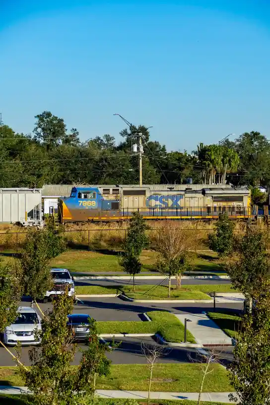 A photograph shows a CSX freight train, locomotive number 7868, passing through a suburban area with a parking lot and trees…