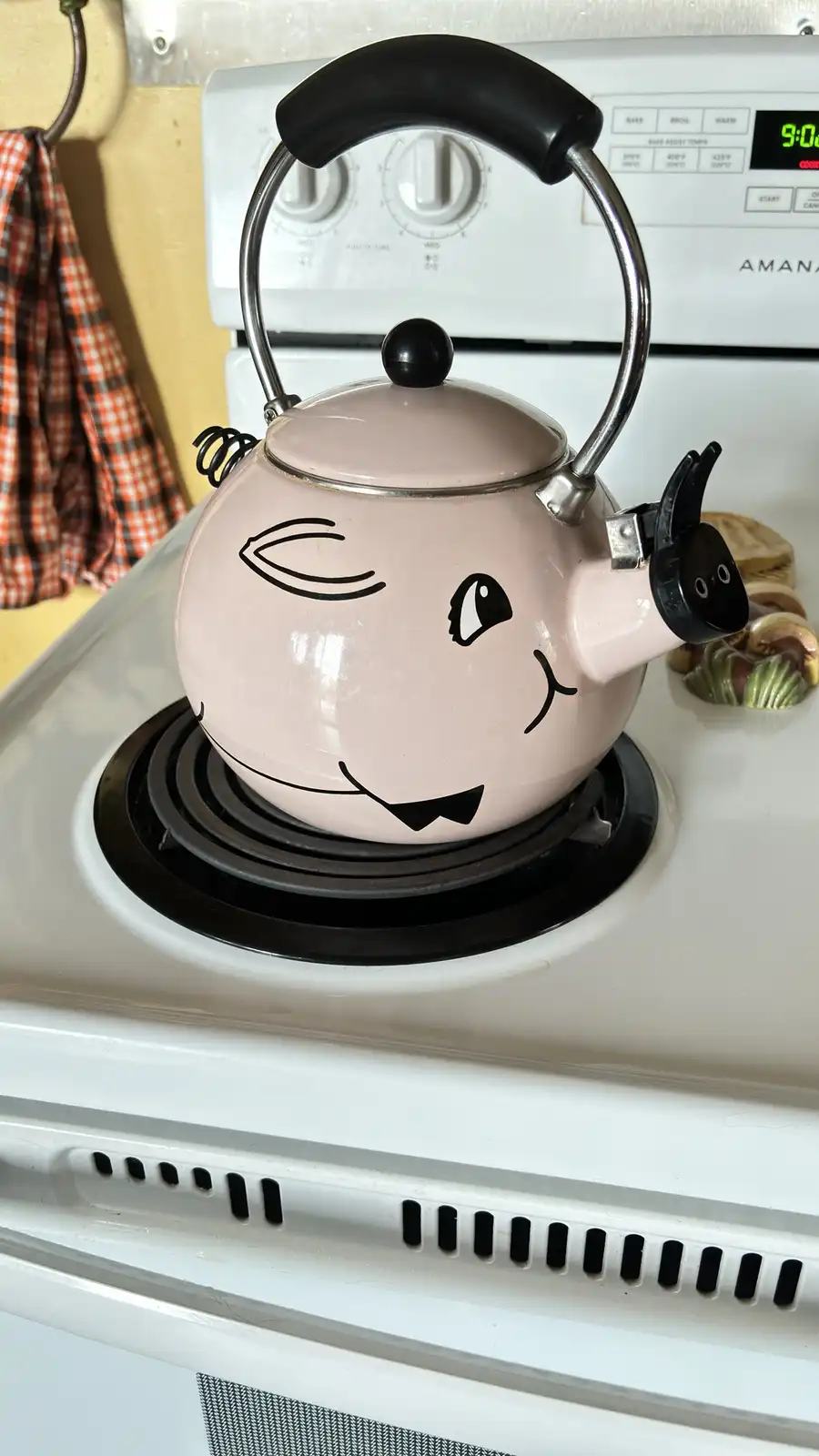 A pink kettle with a hand-drawn cartoon face, featuring one eye open and one eye closed with a smiling mouth, sits on a…