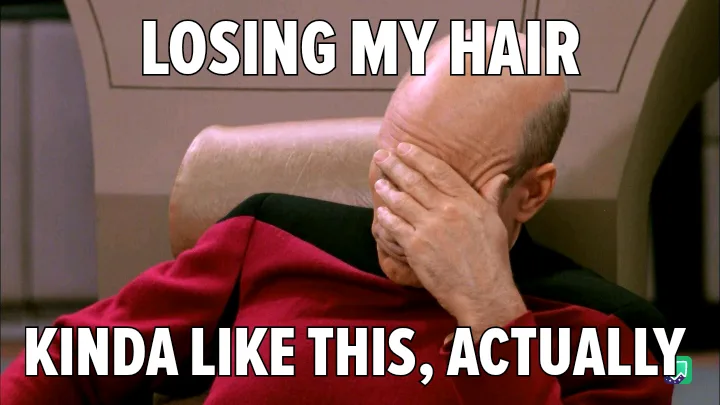 A bald man in a red Starfleet uniform sits in a chair, covering his face with his hand in a gesture of despair or…