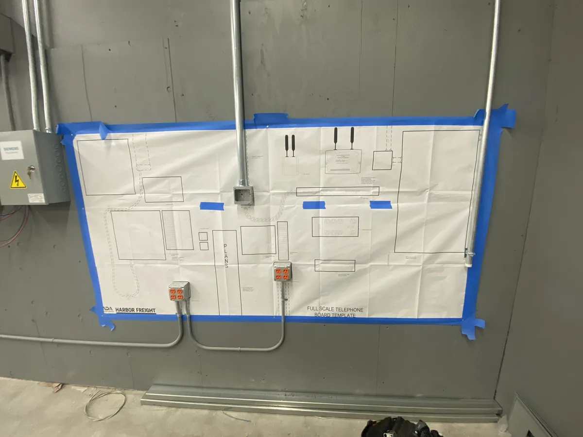 A large blueprint labeled 'FULL SCALE TELEPHONE BOARD TEMPLATE' is taped to a gray wall in a construction or utility room…