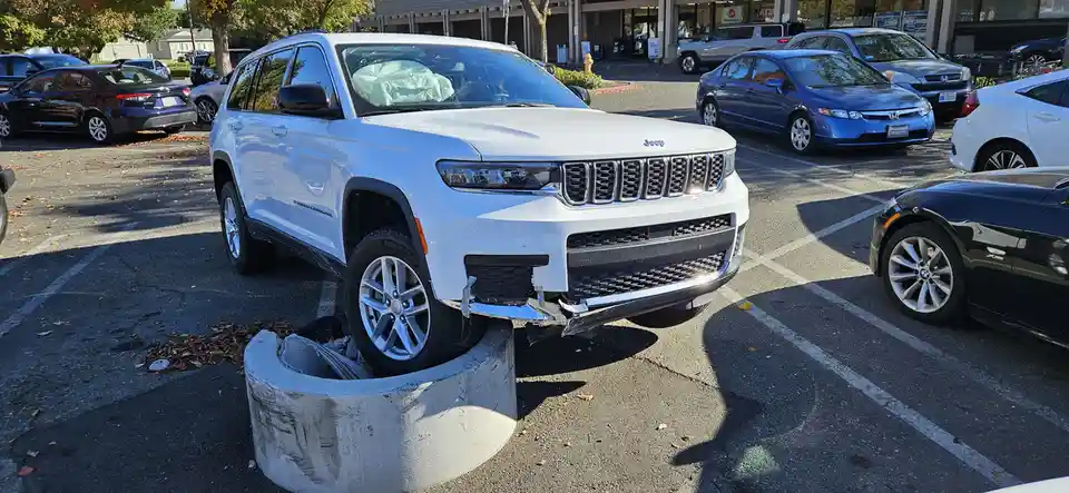 A white Jeep Grand Cherokee is visibly stuck with its front left wheel and bumper lodged into a concrete parking barrier in…