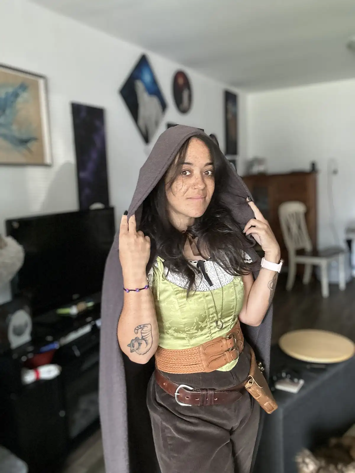 A woman in a homemade fantasy costume, including a grey hooded cloak, green corset, and brown belts, poses indoors while…