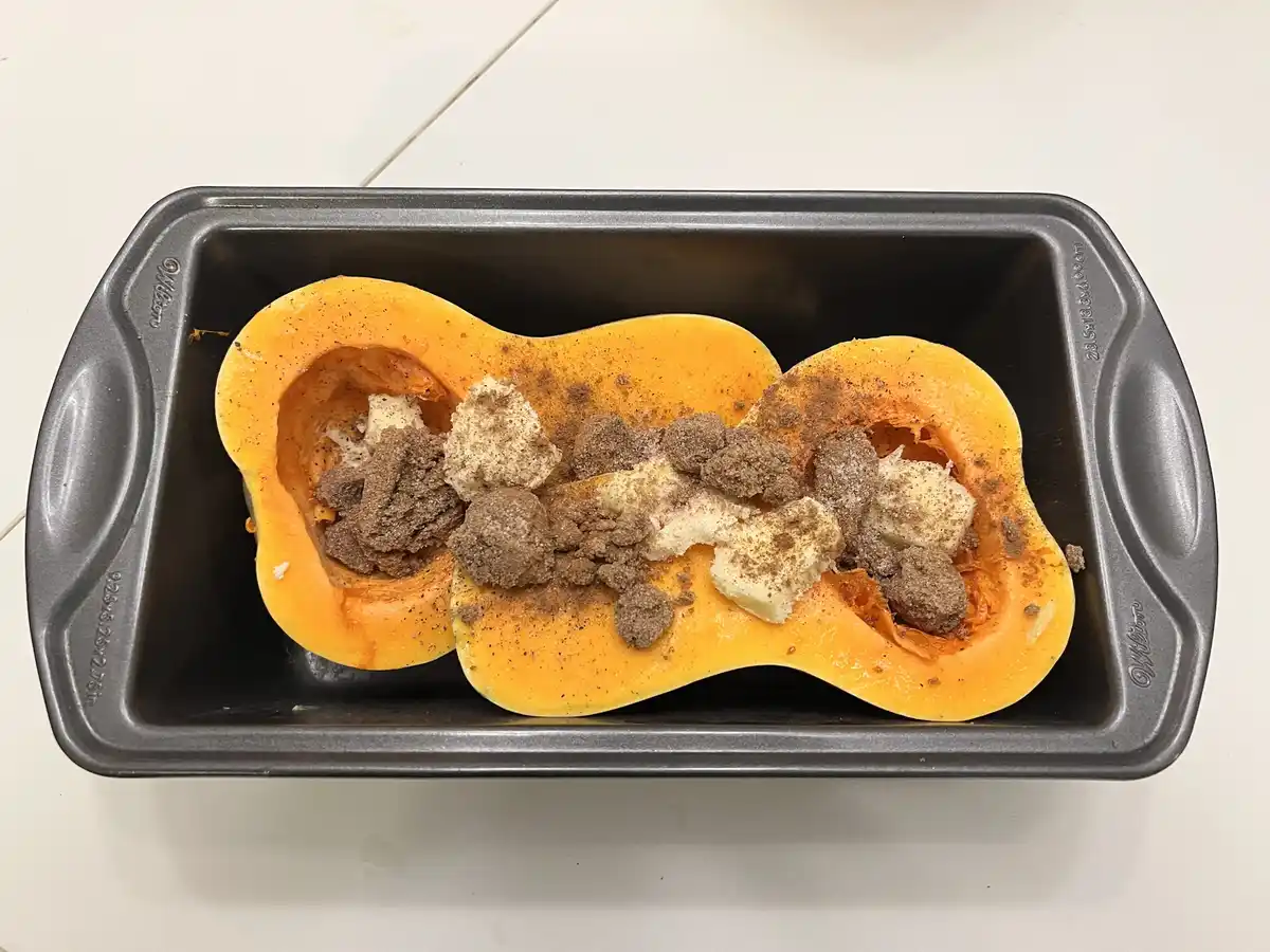 A halved butternut squash sits in a loaf pan, filled with butter, brown sugar, and spices, visually resembling a pumpkin…