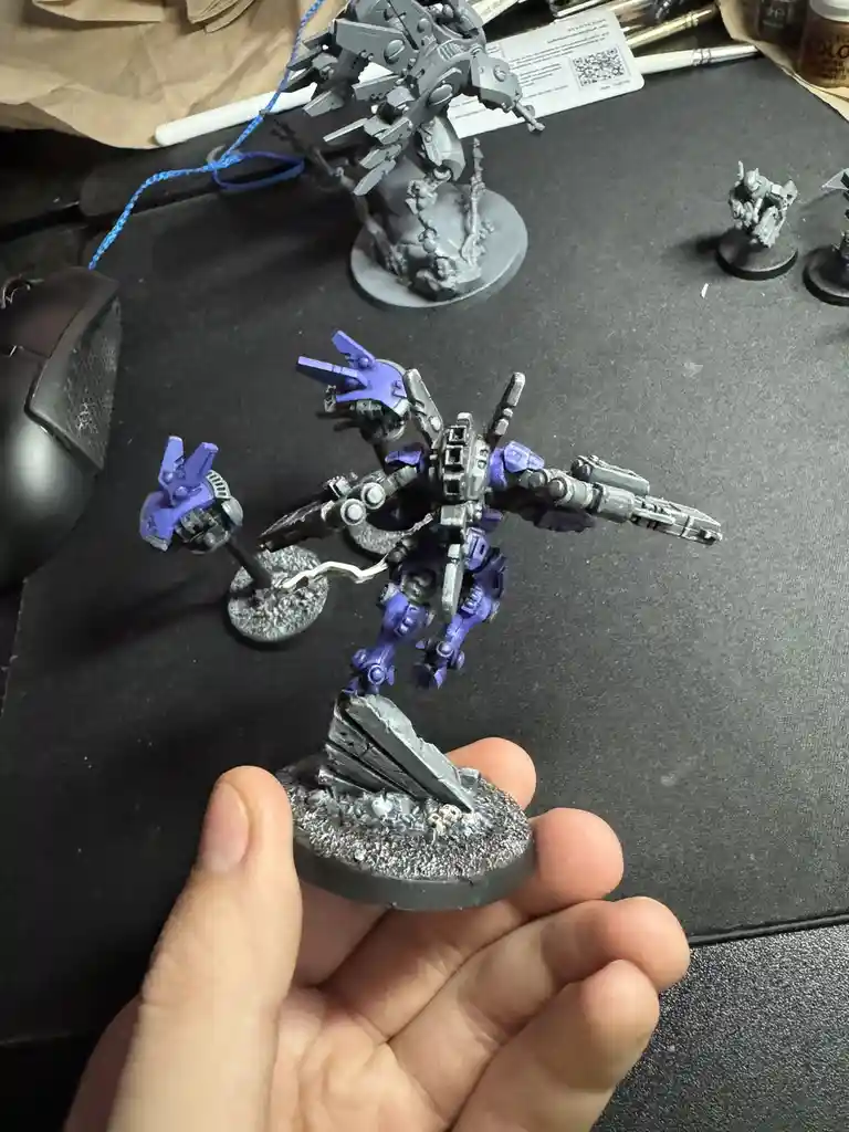 A hand holds a painted miniature mech with purple accents, standing on a rocky base, surrounded by other unpainted mechs and…