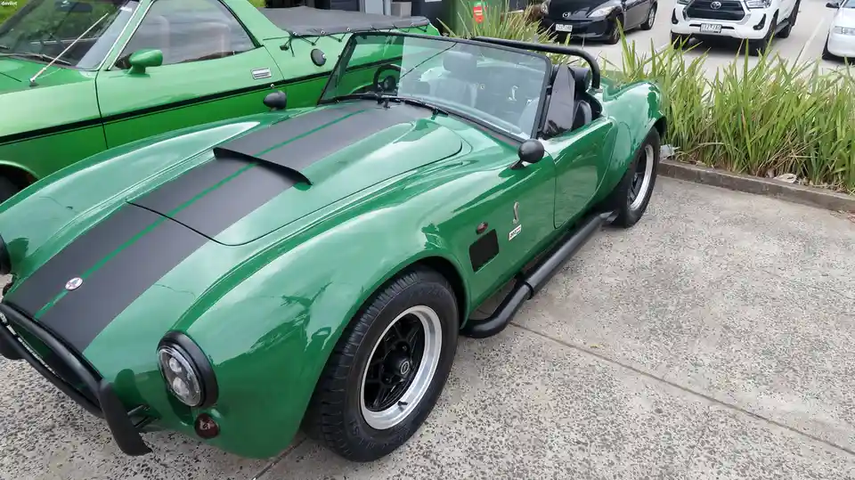 A shiny green classic convertible sports car with black racing stripes and exposed exhaust pipes is parked in a lot, with…
