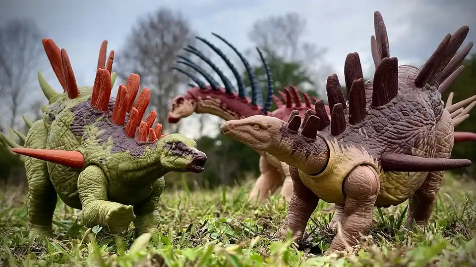 Three plastic dinosaur toys are staged outdoors on grass under an overcast sky. A green stegosaurus with orange spikes is…