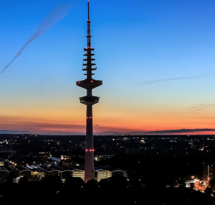 A tall, slender tower with red lights stands silhouetted against a twilight sky with orange and blue gradients, overlooking…