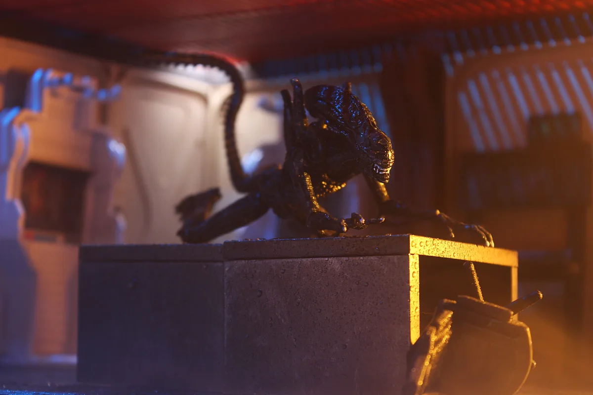 A black alien action figure is posed crouching on top of a metal box in a dimly lit, sci-fi themed setting with orange and…