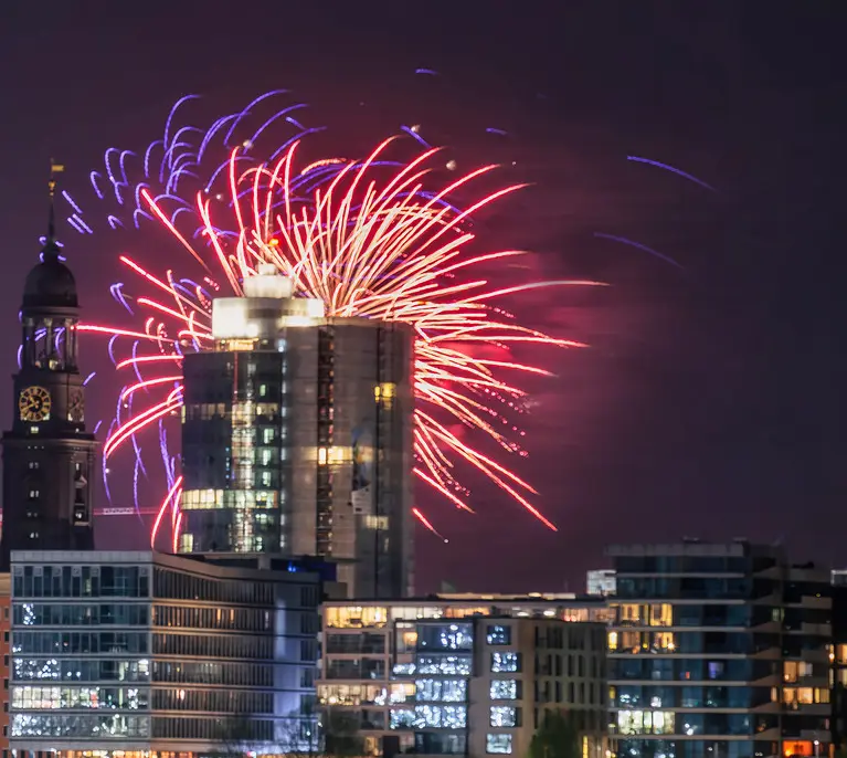 A nighttime cityscape featuring a large building with fireworks exploding directly in front of it, illuminating the scene…