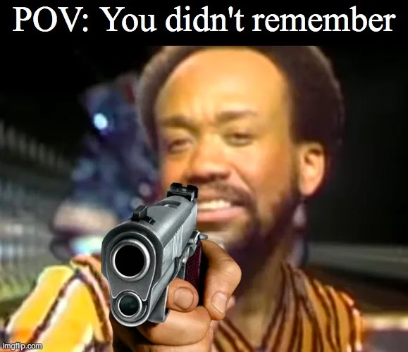 A man with a beard and afro hairstyle holds a silver handgun pointed directly at the viewer. The image has white text at the…