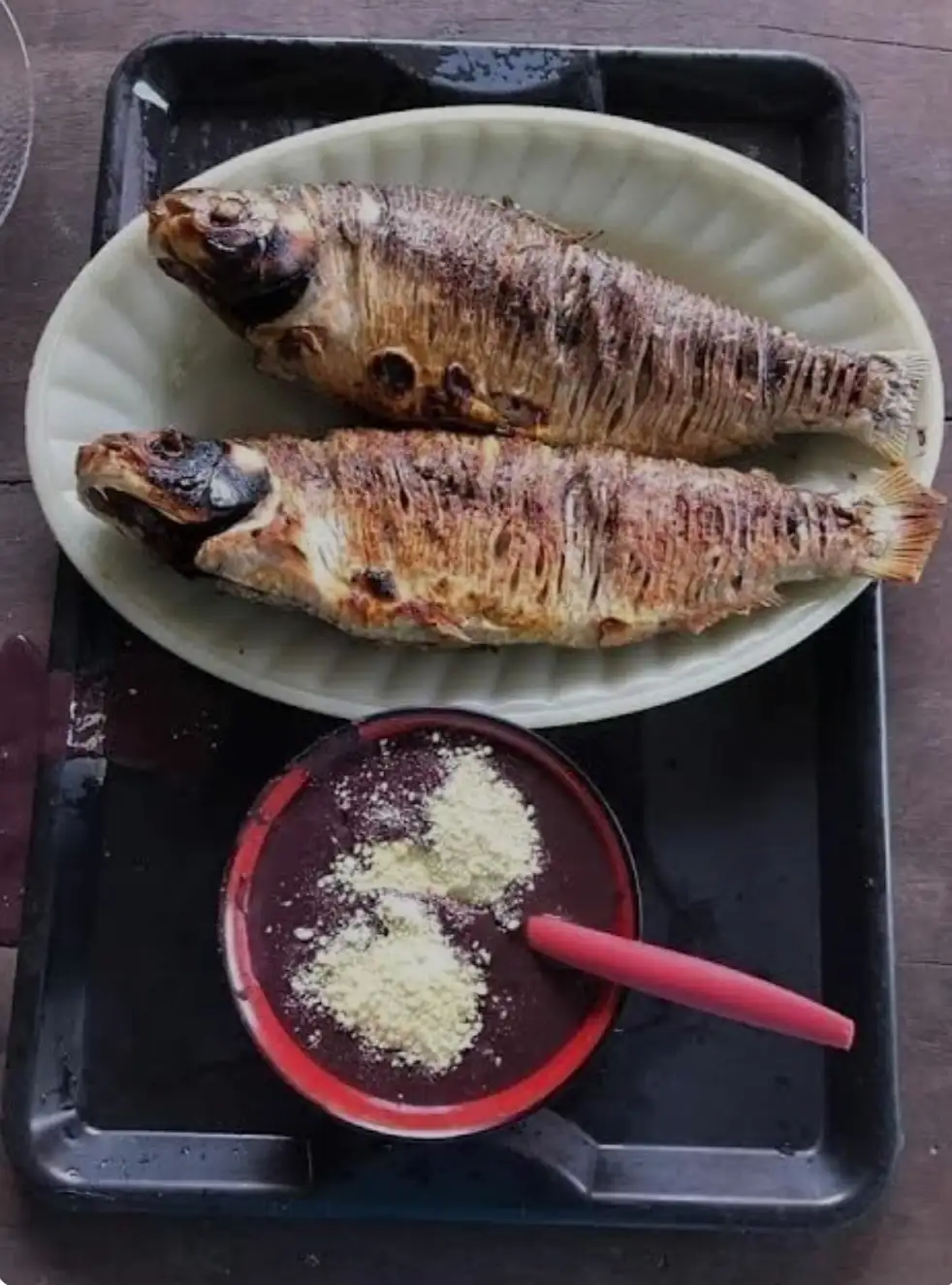 Two grilled fish sit on a white plate atop a black tray, next to a bowl of dark purple liquid with a pink spoon and a…