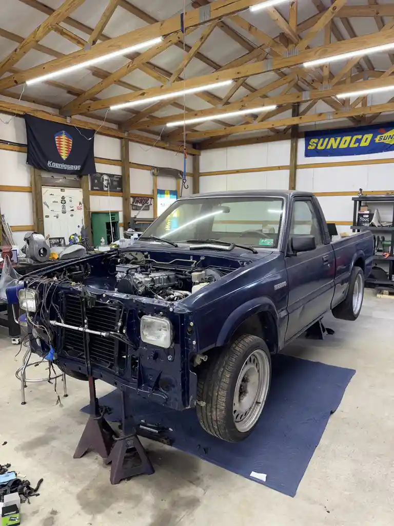 A blue pickup truck is shown in a garage with its hood open and front end partially disassembled, resting on jack stands…