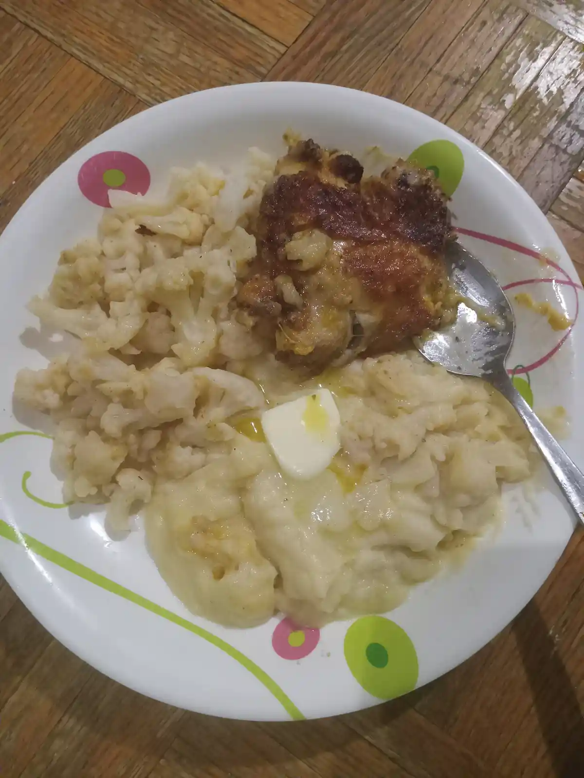 A plate of food featuring a piece of roasted chicken, mashed cauliflower, and a pat of butter melting on top, with a spoon…