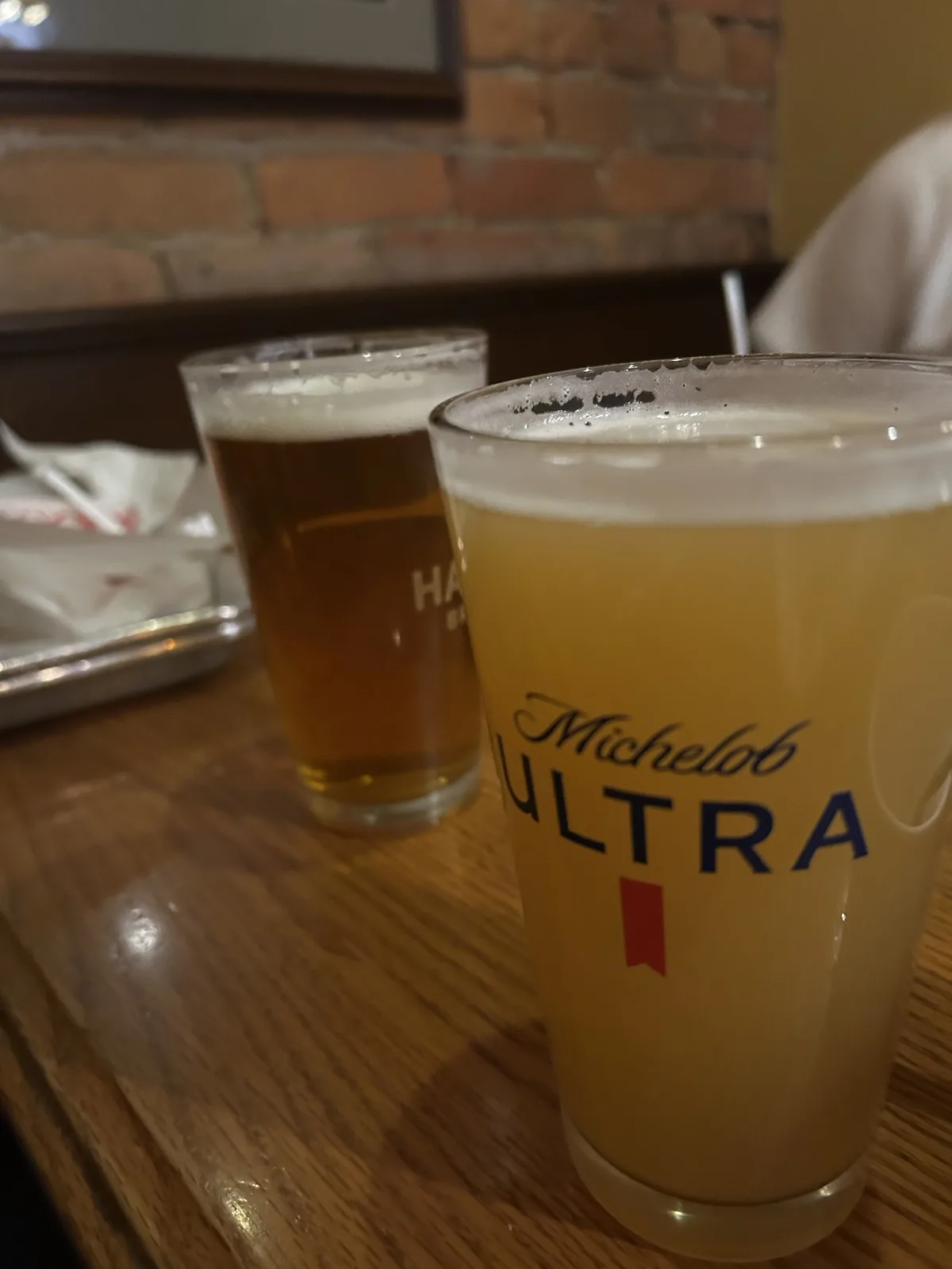 Two glasses of beer sit on a wooden table in a restaurant with a brick wall background; the foreground glass clearly shows…