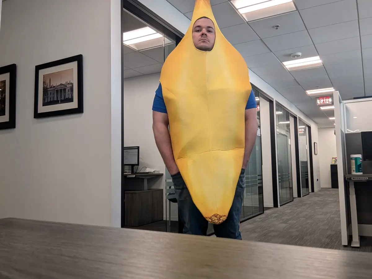 A man stands in an office hallway wearing a full-body banana costume, with only his face visible through the top opening. He…