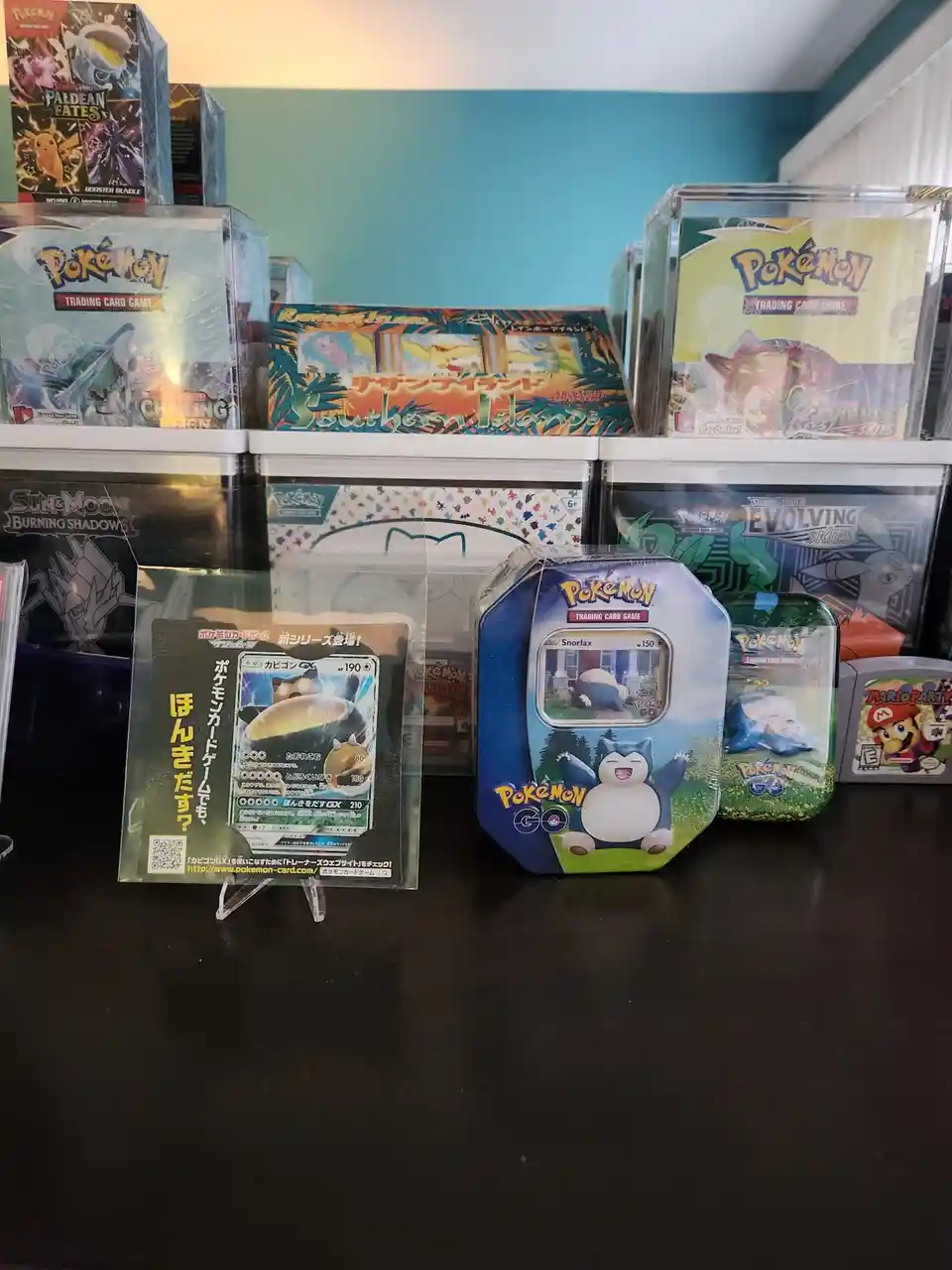 A collection of Pokémon trading card game products, including booster boxes, tins, and a card display stand, arranged on…