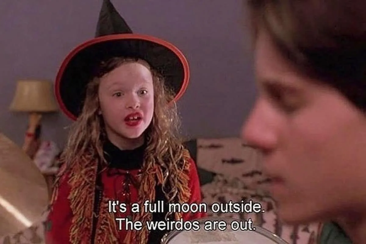 A young girl in a witch costume with a black pointed hat and red outfit stares with wide eyes and an open mouth at a blurred…
