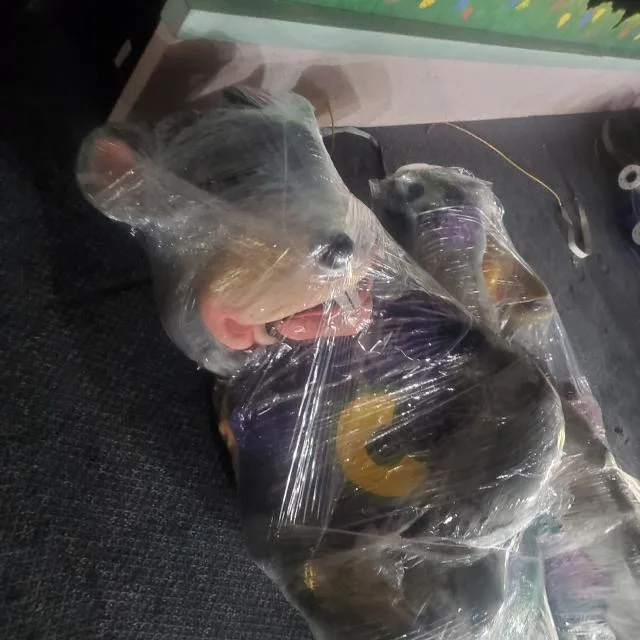 A plush monkey toy, wearing a dark blue shirt with a yellow 'C' logo, is entirely wrapped in clear plastic wrap, lying on a…