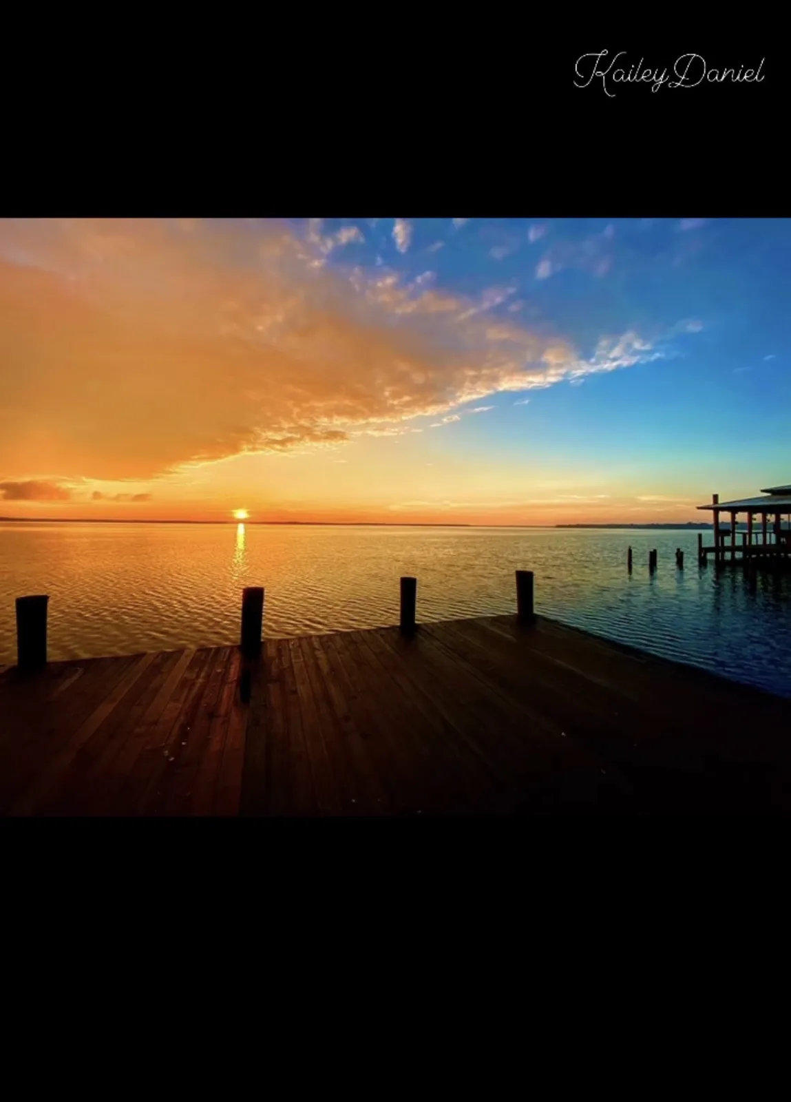 A serene sunset photo shows a wooden dock extending over calm water, with the sun setting on the horizon and orange and blue…