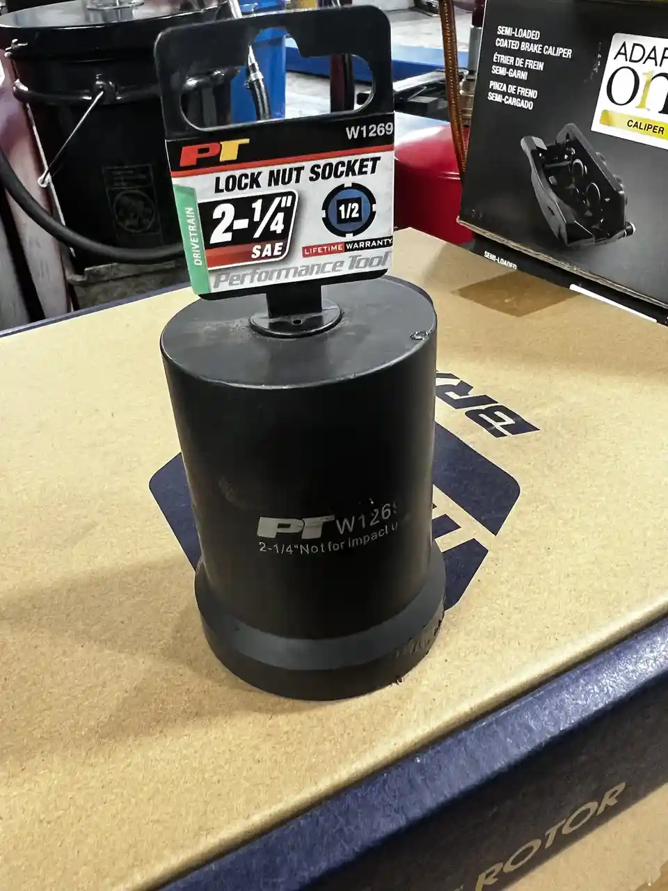 A black PT brand lock nut socket, model W1269, size 2-1/4" SAE, sits on a cardboard box labeled 'ROTOR'. The tool's…