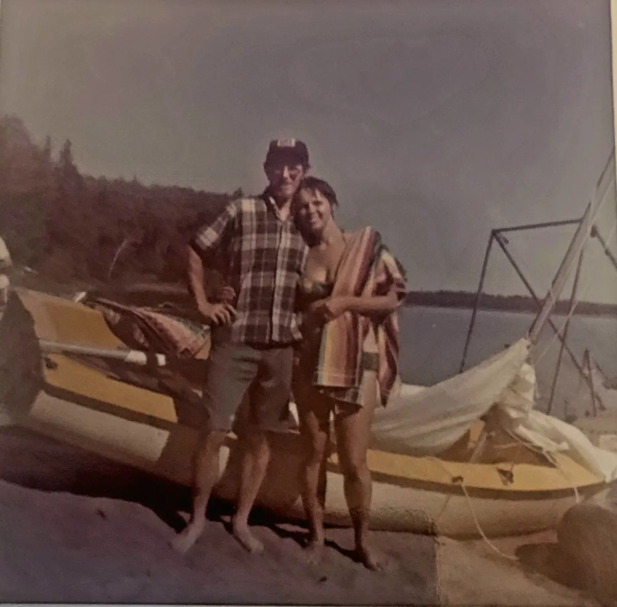 A faded, vintage-style photo shows a man and woman standing barefoot on a dock next to a yellow sailboat with its sail down…