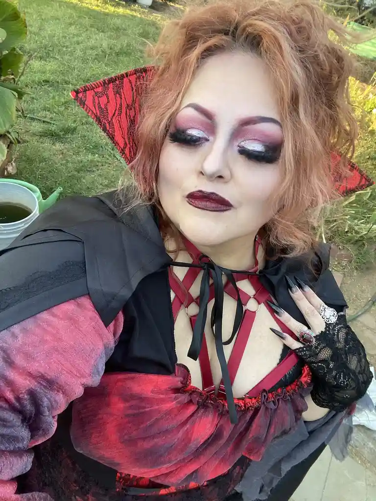 A person with red hair and dramatic makeup, wearing a black and red gothic outfit with pointed red ears, poses outdoors…