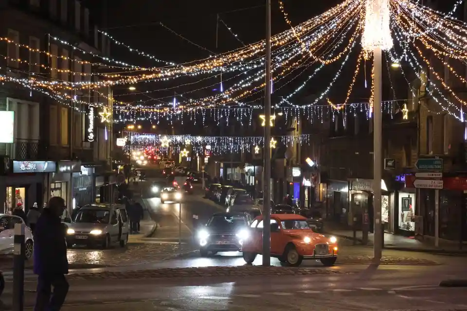 A nighttime street scene decorated with festive string lights and star-shaped ornaments, featuring cars and pedestrians. An…