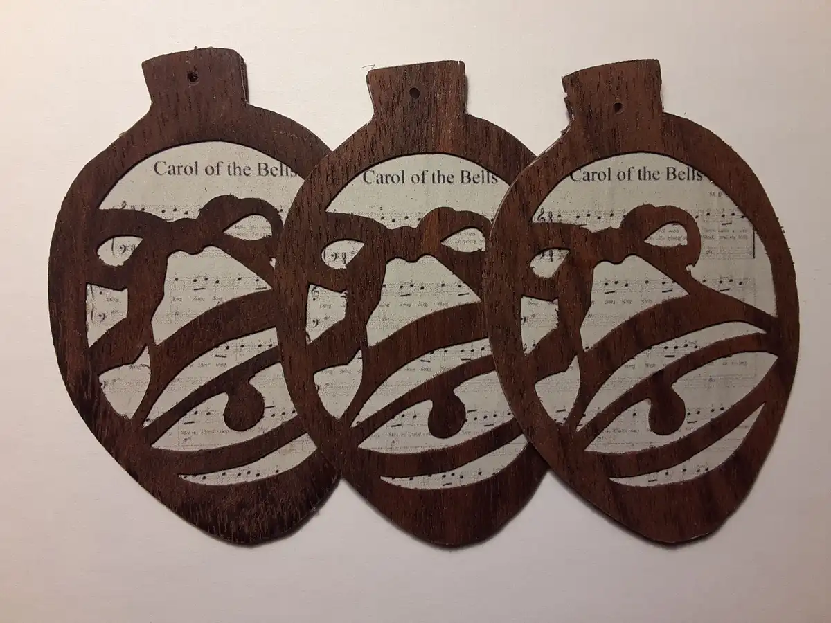 Three identical wooden ornaments shaped like bells, each with a cutout design revealing sheet music underneath. The text…