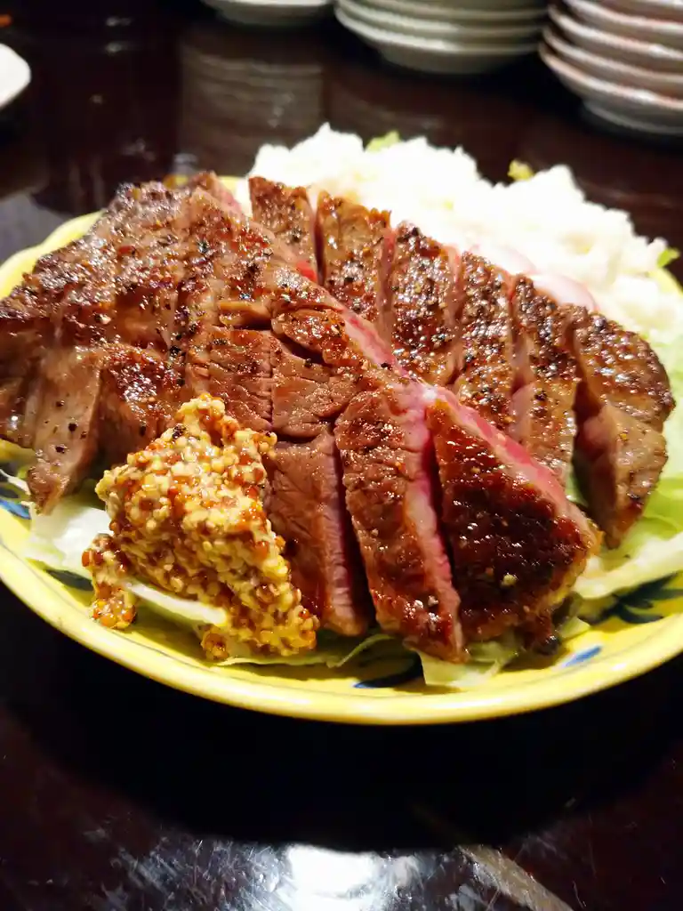 A close-up photo shows a plate of sliced, medium-rare steak with a seared crust, served over lettuce and rice with a side of…