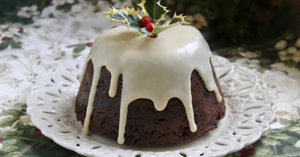 A close-up of a traditional dark Christmas pudding with creamy white icing dripping down its sides, topped with a holly…