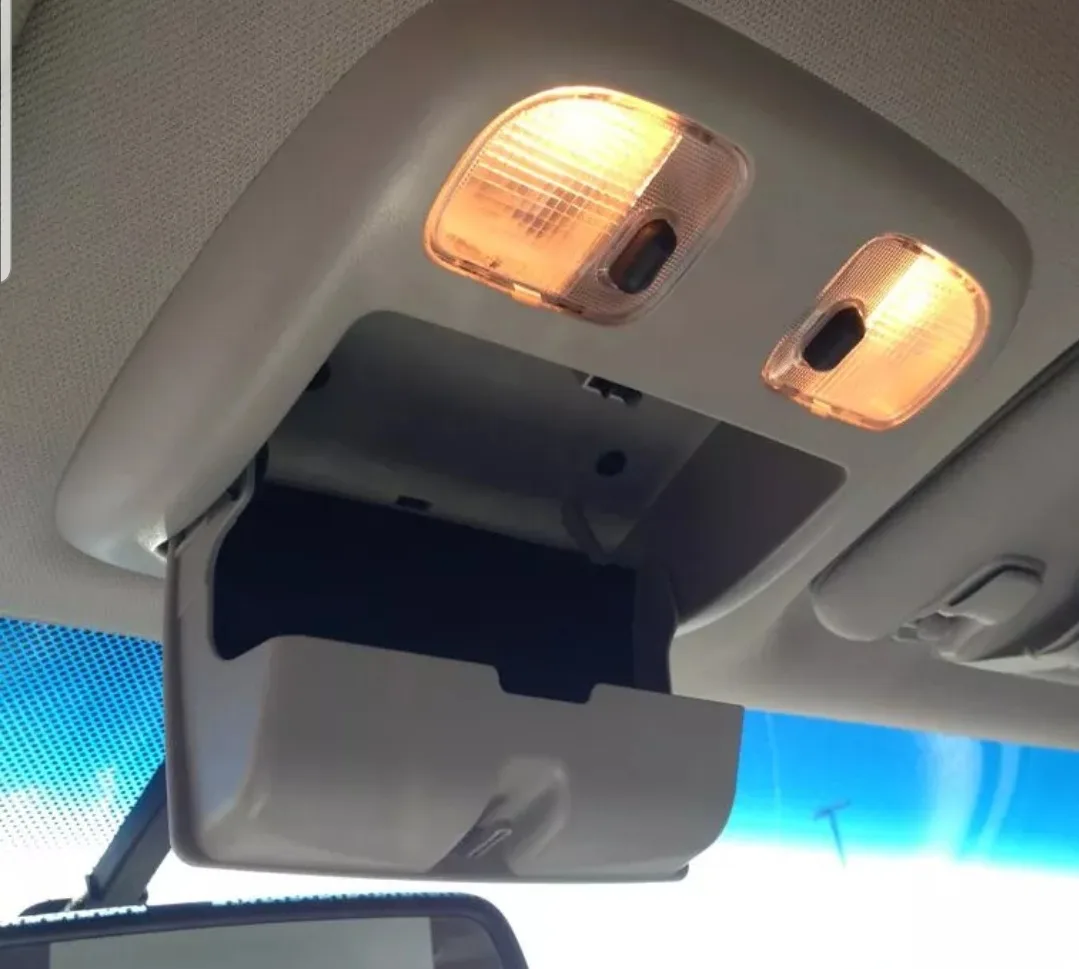 A close-up photo of a car's interior ceiling, showing two illuminated amber dome lights above an open sun visor. The visor…