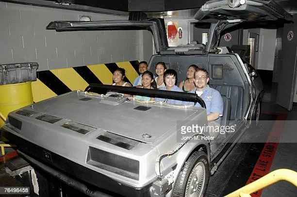 A group of smiling people are seated inside a silver DeLorean time machine, likely at a theme park ride, with its gull-wing…