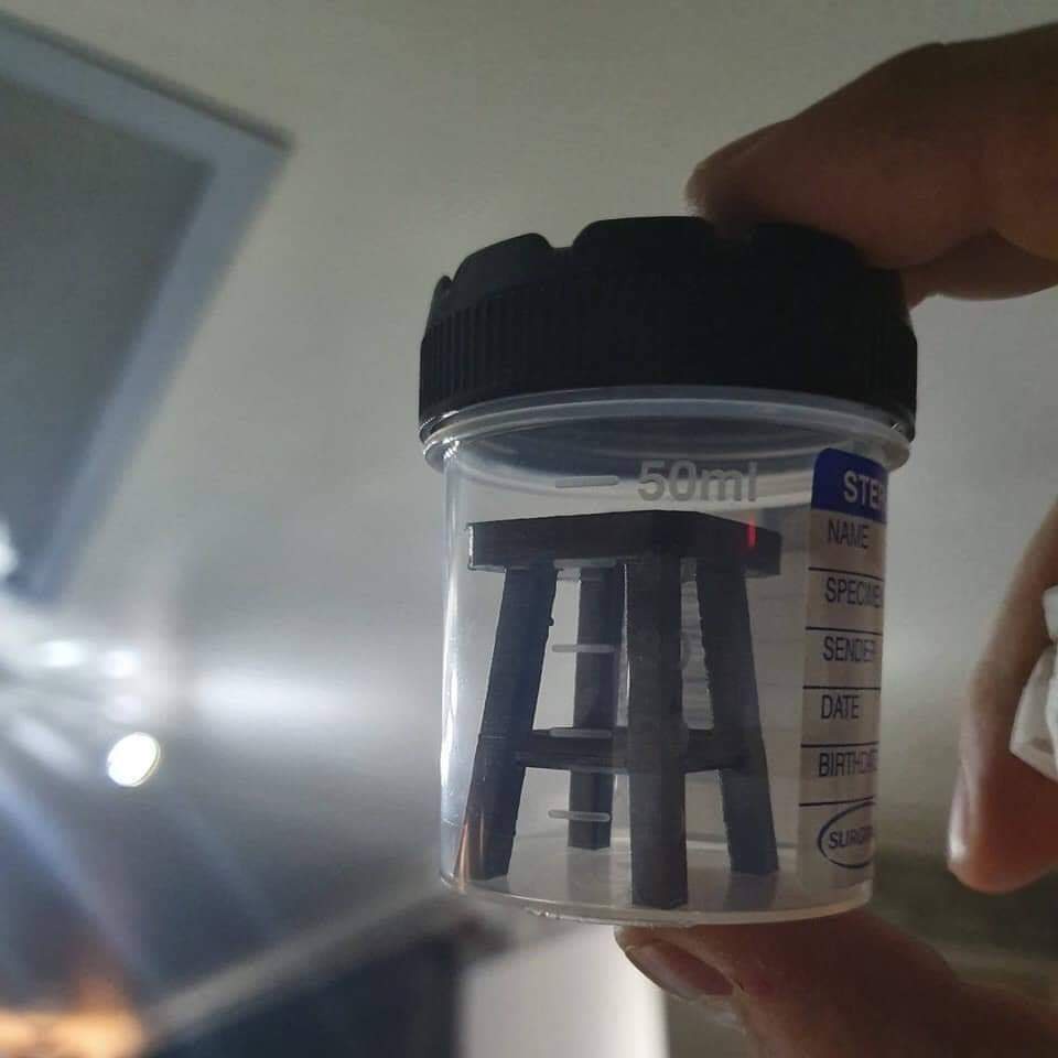A hand holds a clear 50ml medical specimen container with a black lid, inside which sits a miniature black stool. The…