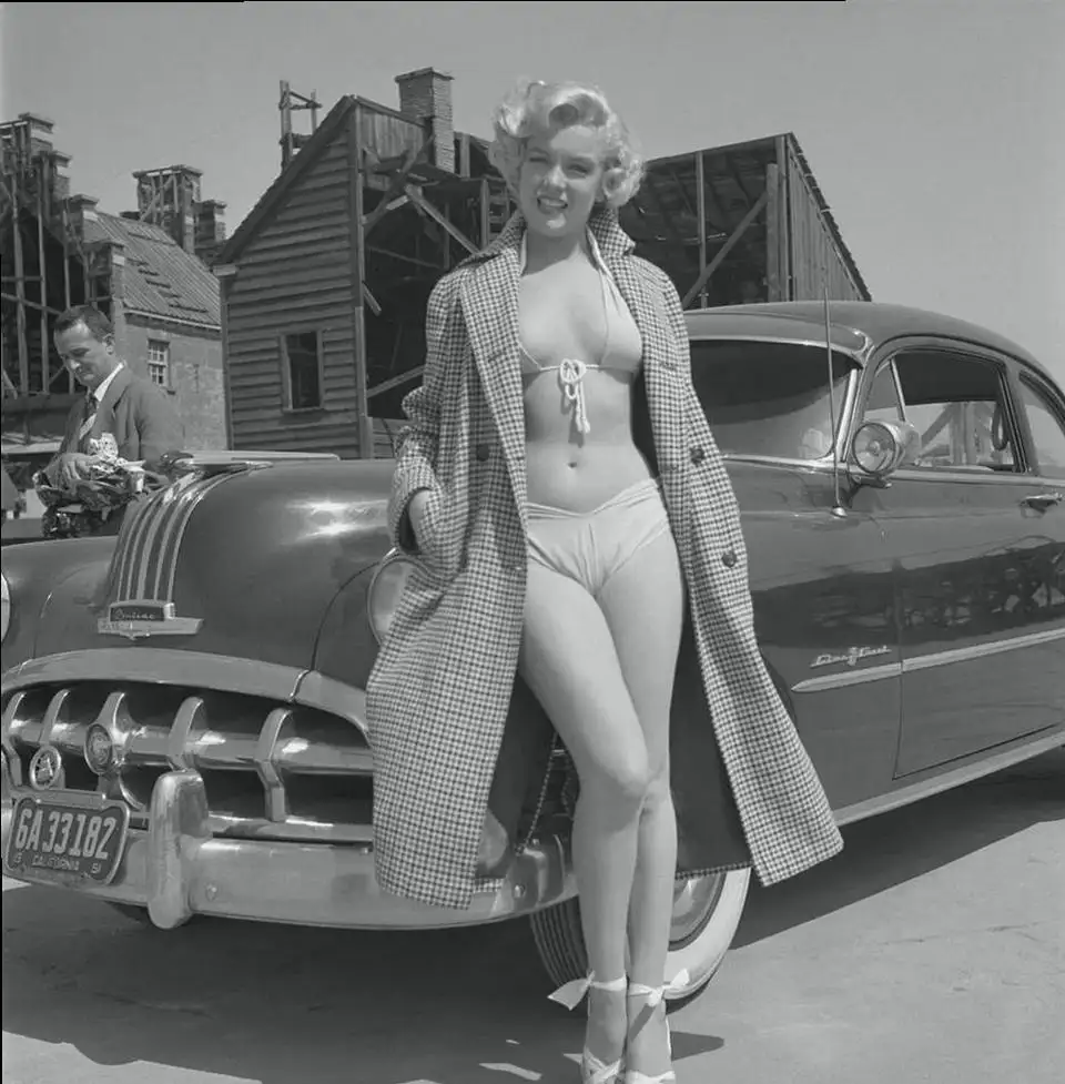 A black-and-white photo shows Marilyn Monroe in a bikini and checkered coat leaning against a vintage car, with a man in the…
