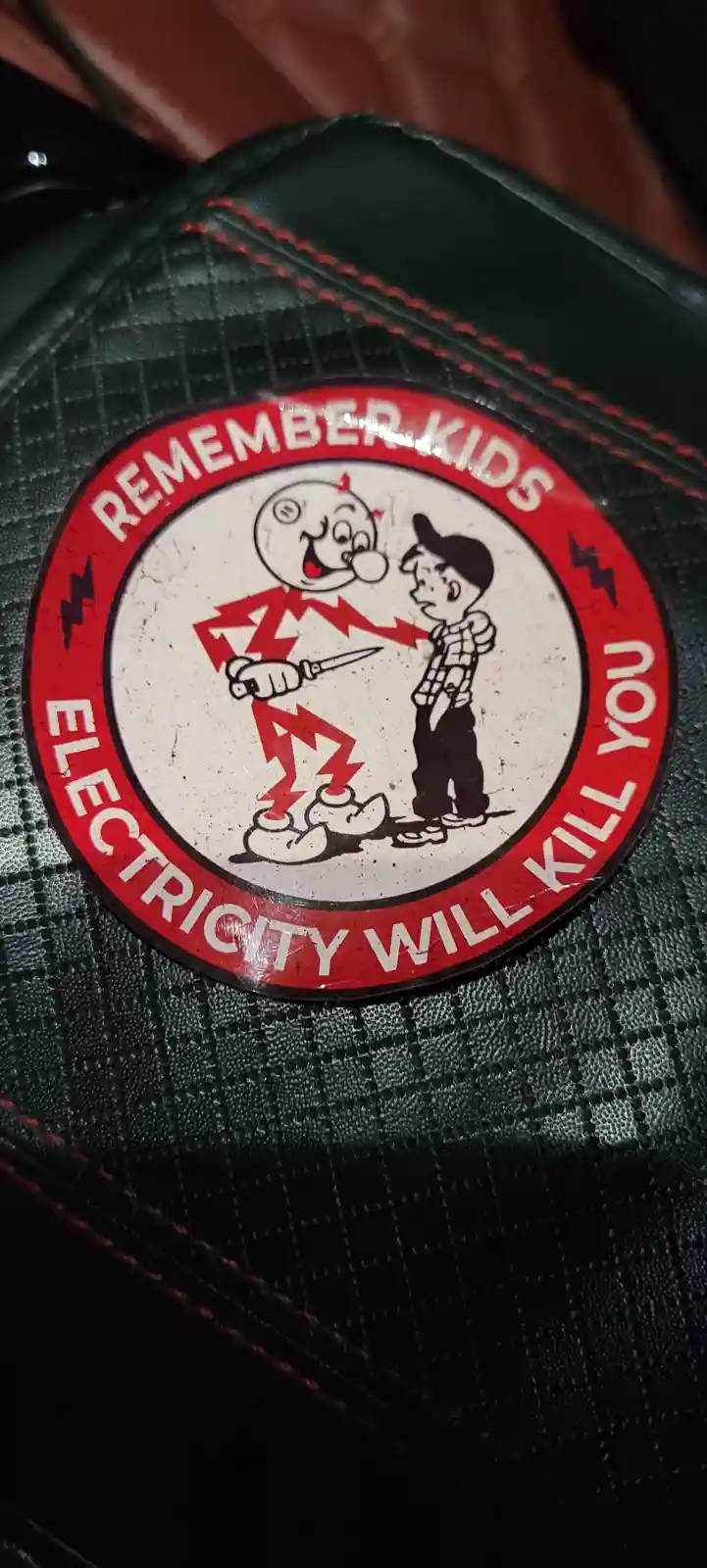A circular sticker with a red border and white center shows a cartoon electric current character holding a knife and smiling…