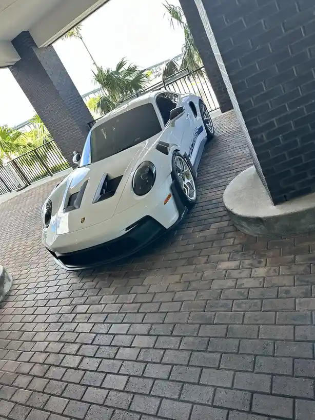 A white Porsche 911 GT3 RS sports car is parked at an angle on a brick driveway under a covered entryway, with palm trees…