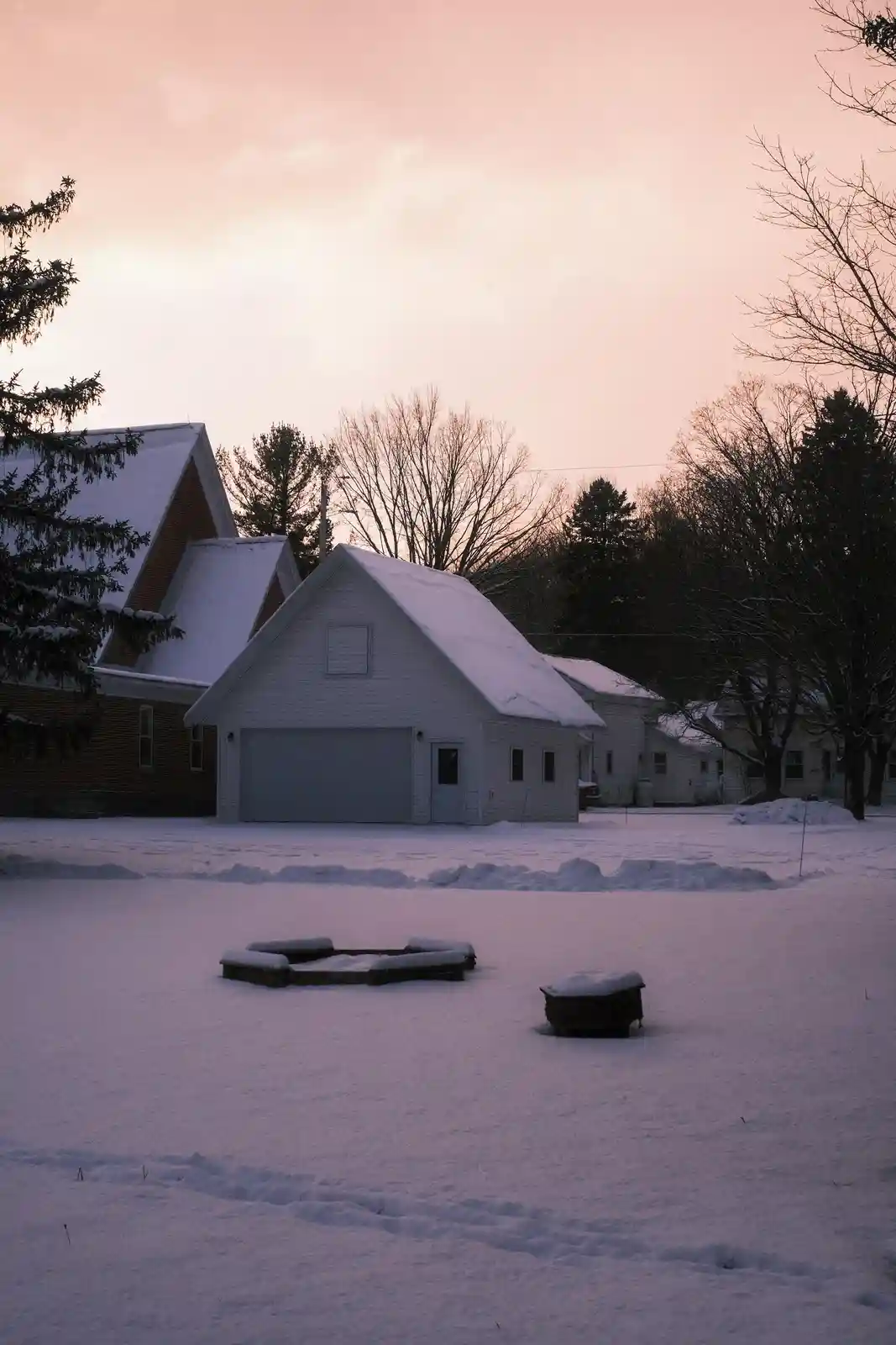 A snowy suburban yard at dusk shows a white garage and brick house with snow-covered roofs and bare trees. In the…