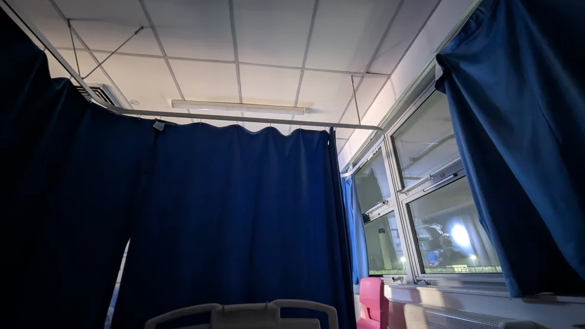 A low-angle shot from inside a hospital room, showing blue privacy curtains, a hospital bed headboard, and a pink plastic…
