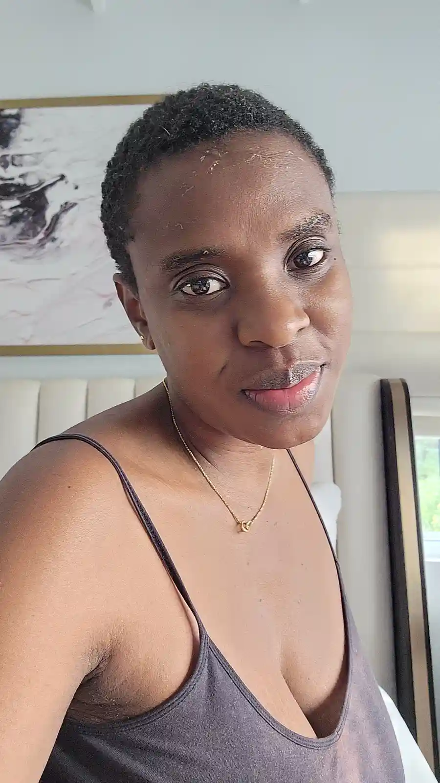 A person with short dark hair and a grey tank top looks at the camera with a slightly amused expression, their face and lips…