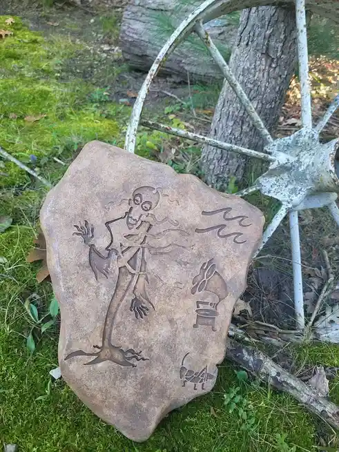 A carved stone in a mossy outdoor setting depicts a cartoonish skeleton figure with wild hair, arms raised, next to a…