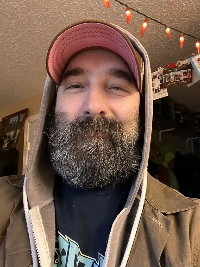 A man with a full gray beard and mustache wears a pink and brown baseball cap backwards under a gray hoodie, smiling…