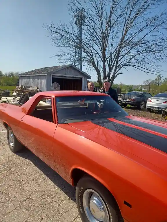 Two men stand behind a bright orange classic muscle car with black racing stripes, smiling at the camera. The car, likely a…