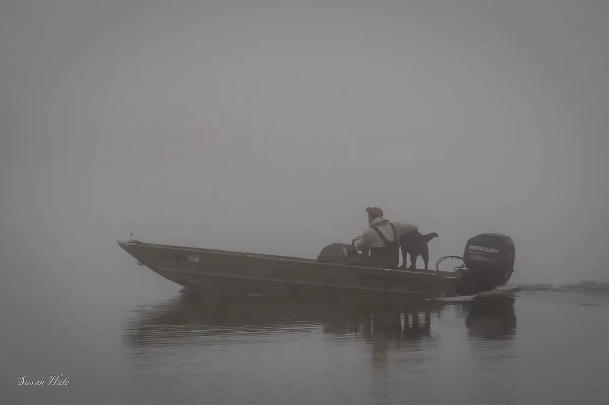 A person and a dog are in a small boat on a foggy lake, with the boat's motor visible. The image is enveloped in thick fog…