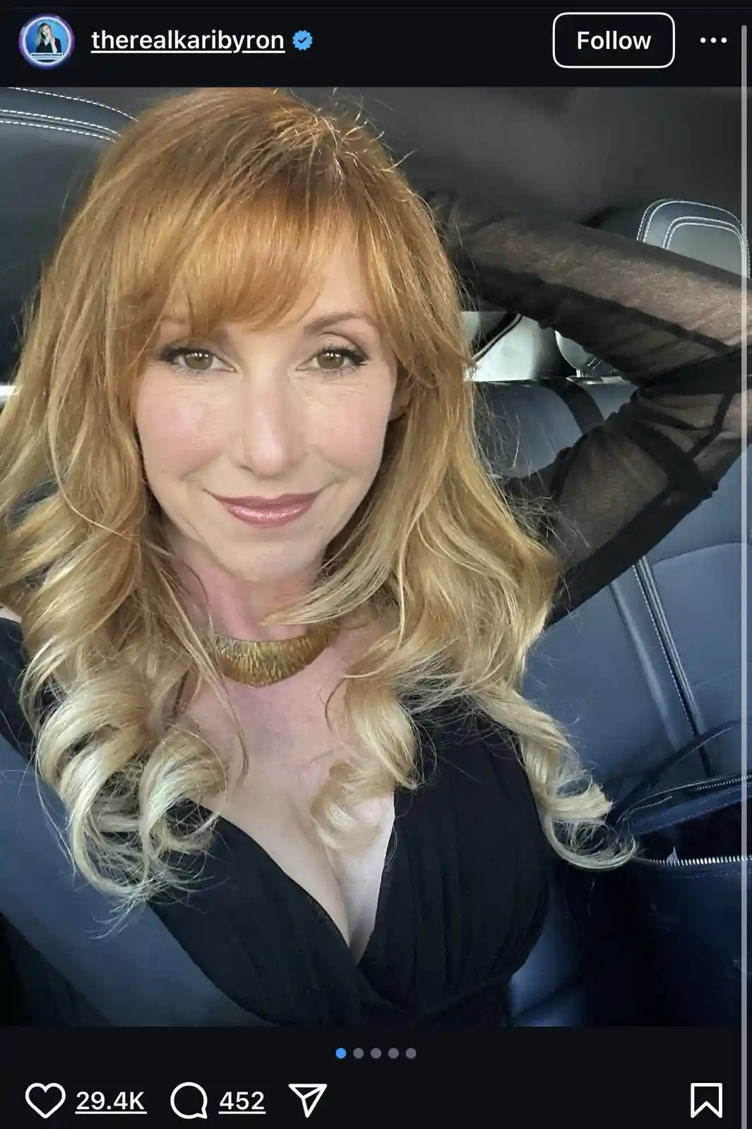 A woman with blonde hair and a gold choker sits in a car wearing a black top, smiling at the camera. The image is framed as…