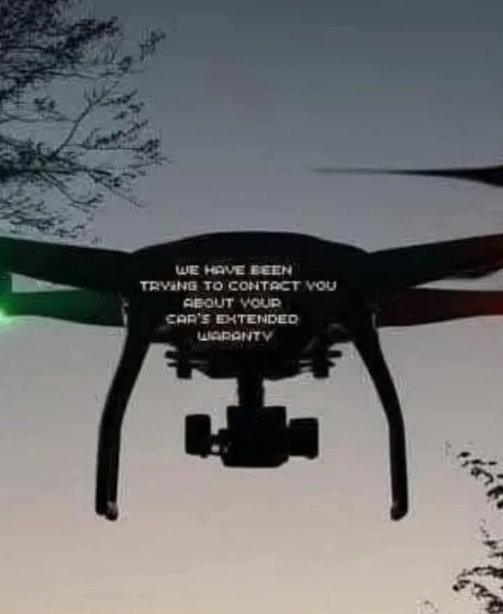 A black drone is shown flying against a twilight sky with tree branches visible at the top and bottom. The drone's body has…